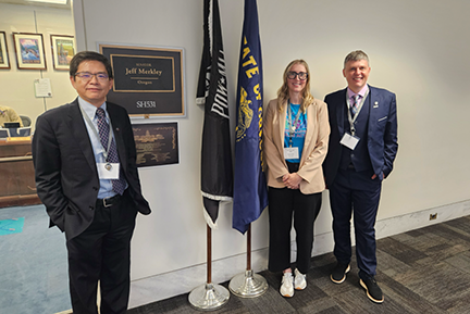 Two men and one woman stand in a hall outside of a staffer's office. There are two flags on flag poles between them.