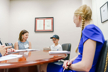 A young boy affected by ectodermal dysplasia is wearing glasses and a baseball hat. He's reading from a notebook his story. He sits at a conference table next to his mom while a legislative staffer listens,