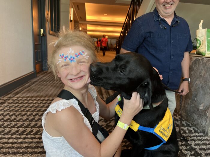 A young girl is holding a black lab who is licking her face. She's smiling and laughing.