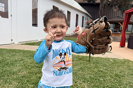 A young boys has his arm in an adult size baseball glove and  he's holding up one finger.