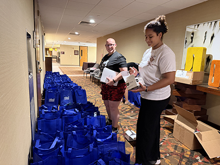 A woman who is bald and wearing glasses and a tween girl are holding items to put into Family Conference goodie bags.