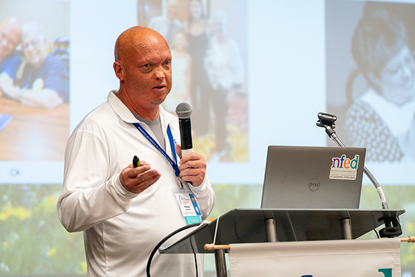 A man stands at a podium talking into a microphone. A slide of photos shows behind him.