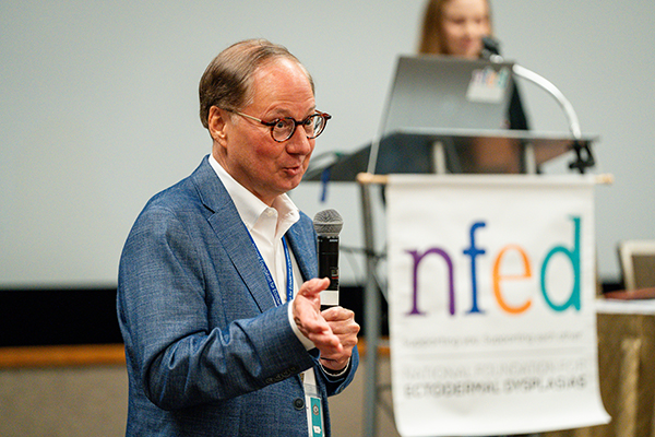 A man is talking into a microphone speaking to a crowd. A woman is in the background behind a podium. An NFED banner hangs from the podium.