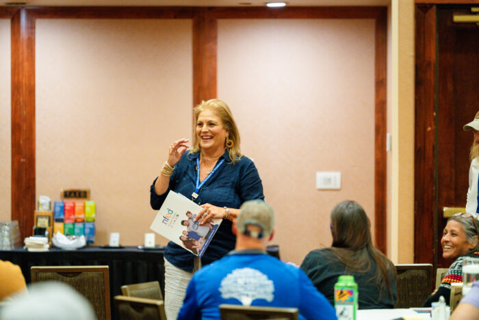 A woman stands in front of a group of people in a conference room. She's holding an NFED folder and is smiling.