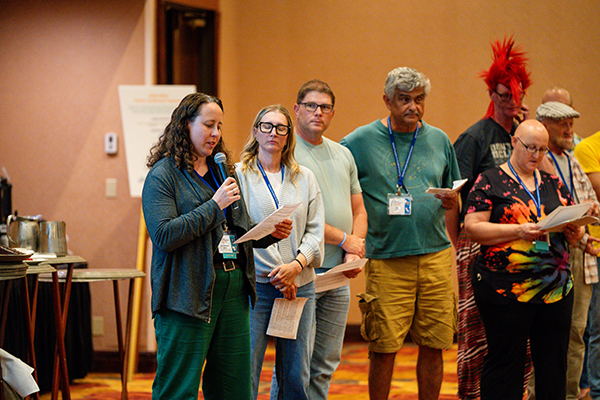 A woman is speaking into a microphone, reading a piece of paper. To her right, numerous people are standing getting ready to read their parts.