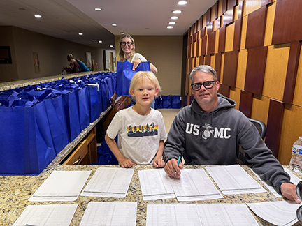 A Family Conference volunteer is sitting at a counter looking at registration sheets with his young son. His wife is behind them lining up Family Conference tote bags for guests.