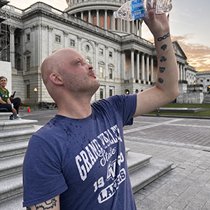 A man affected by ectodermal dysplasia is lifting a bottle of water over his head to pour it on himself to cool off. In the background is the U.S. Capitol Building.