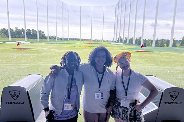 Three young women have their arms around each other and are standing in front of a large golf course.