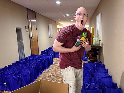 A teenage boy who is bald, wearing a maroon shirt and khaki pants is holding small bags of snacks to put into blue tote bags which are lined up on the floor. He's wearing glasses.