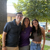 Three adults stand arm in arm outside on a sidewalk with a tree behind them.
