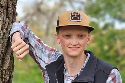 Keegan is standing next to a tree with is forearm on the tree. He's wearing a baseball hat, shirt and vest.