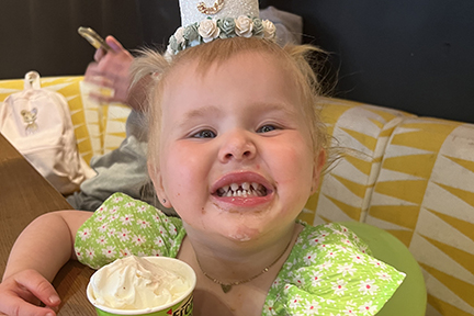A toddler is wearing a green shirt and has small roses in her sparse hair. She's smiling. Her teeth are widely spaced apart and misshapen due to TSPEAR-related ectodermal dysplasia.