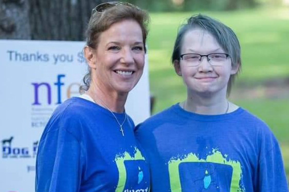 A mom and her son stand shoulder to shoulder wearing matching race tshirts. They are standing outside in front of a sign thanking race supporters.