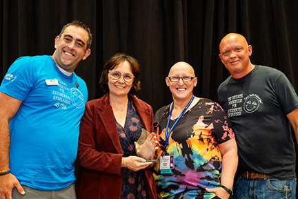 Four people stand on a stage linking arms in front of a black backdrop. A woman is holding an award.