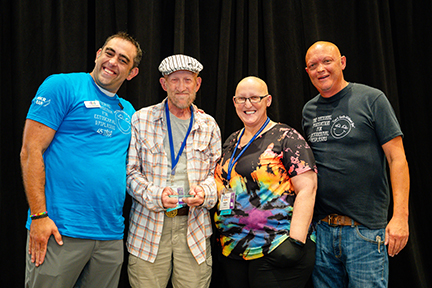 Four people stand on a stage linking arms in front of a black backdrop. A man is holding an award.
