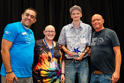 Four people stand on a stage linking arms in front of a black backdrop. A young man is holding an award.