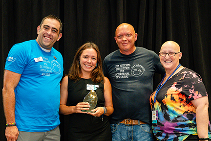Four people stand on a stage linking arms in front of a black backdrop. A woman is holding an award.