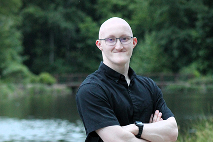 Zane is wearing a black shirt and standing outside with a lake and trees behind him.