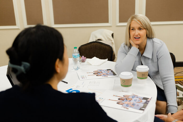 Two women are seated at a round conference table talking.