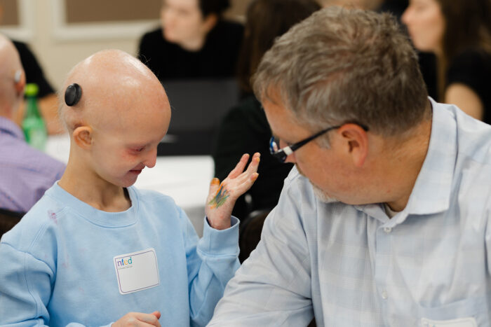 A little girl who is bald and wearing a baja hearing implant holds up her palm to show a doctor how she had colored it with markers.