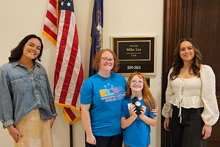 Four people pose in front of a Congressional office with two flags hanging in the background.