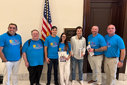 Six advocates in ELSA t-shirts pose with a staffer outside a Congressional office. A U.S. flag hangs behind him.