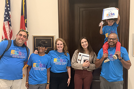 Six people pose for a photo outside a Congressional office. Two flags hang in the background. A child is sitting on his father's shoulders holding a sign that says, "I'm advocating for ELSA."