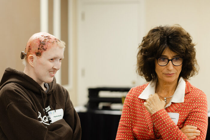 A woman affected by EEC syndrome who has skin erosions on her scalp stands and smiles with a researcher as they listen to someone who is not shown in the picture.